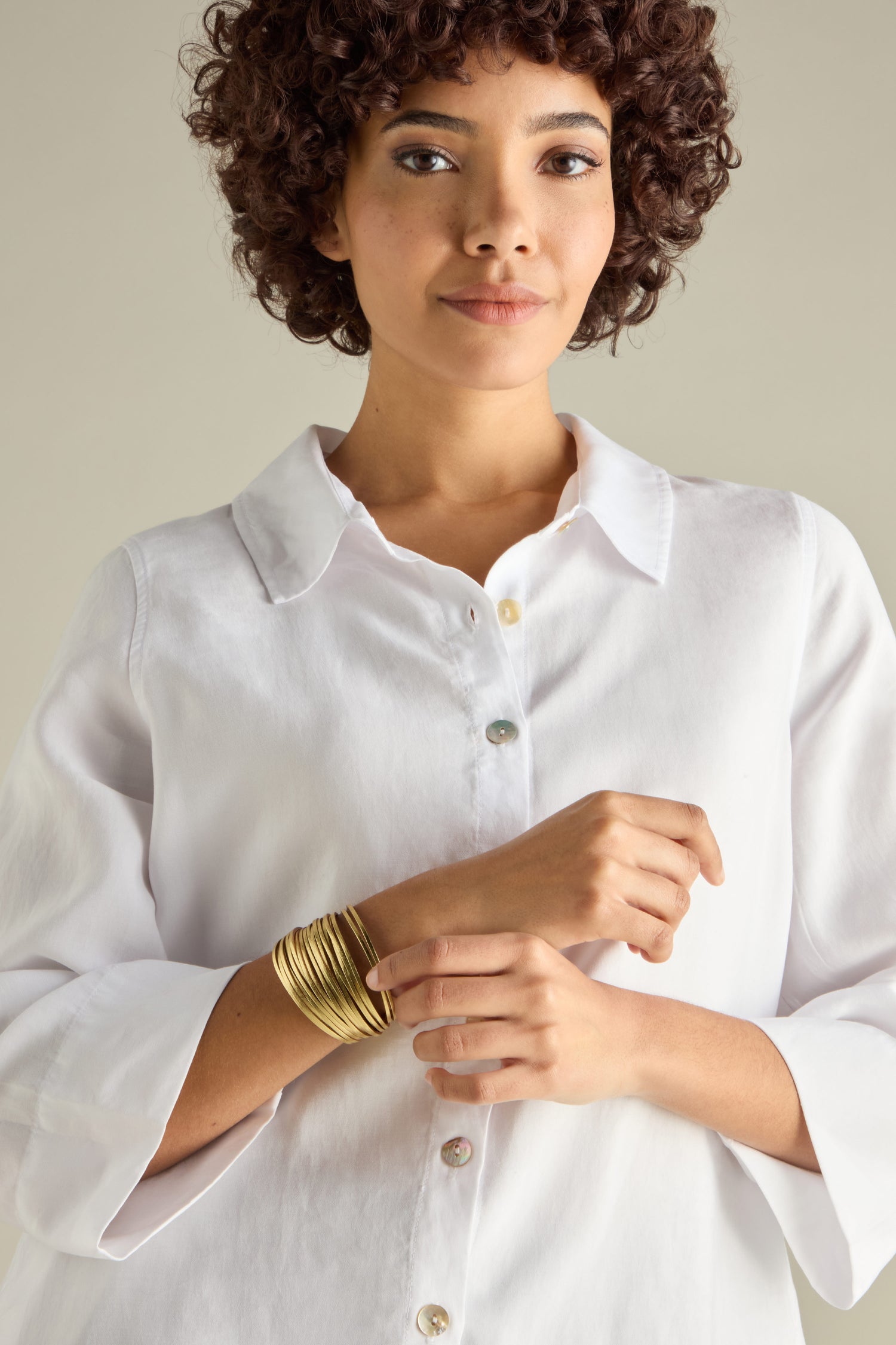 A person with curly hair is wearing a white button-up shirt and multiple gold-colored bangles, one of which is the Woven Metallic Bracelet featuring a magnetic clasp.