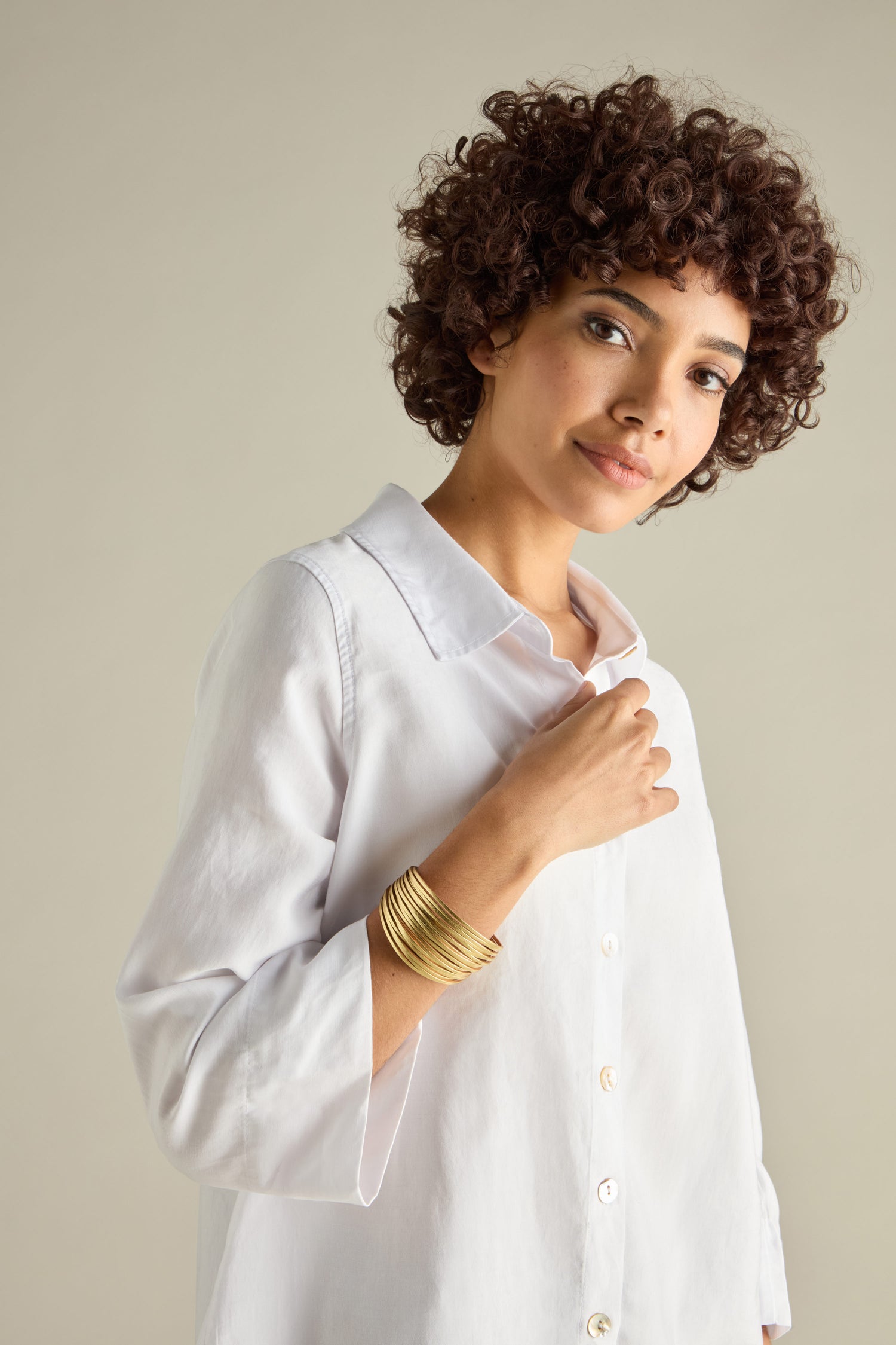 A person with curly hair in a white button-up shirt showcases gold bangles and the Woven Metallic Bracelet, featuring a twisted leatherette design, while gazing confidently at the camera.