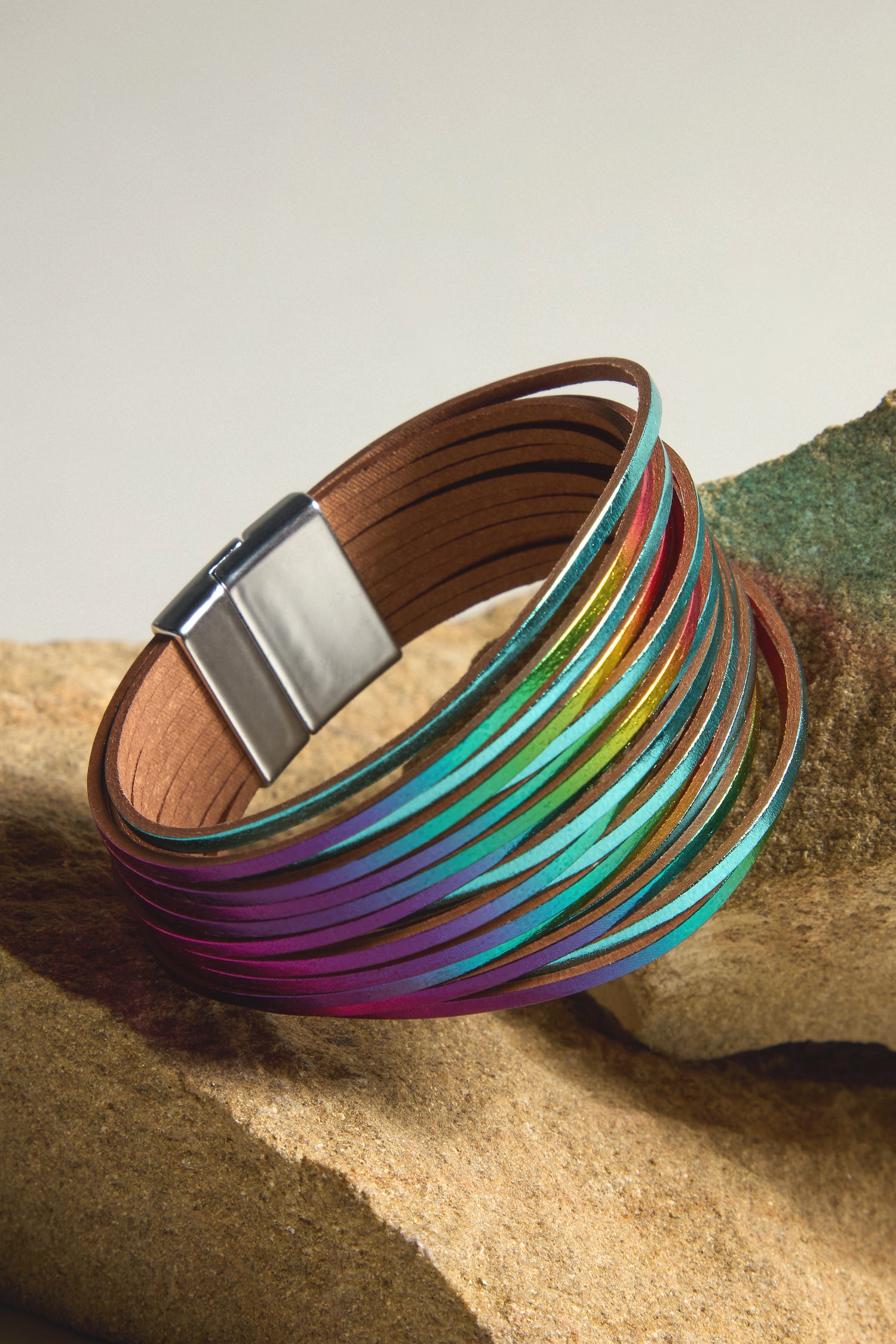 The Woven Metallic Bracelet, featuring a multicolored leather design and magnetic clasp, elegantly rests on a textured stone surface.