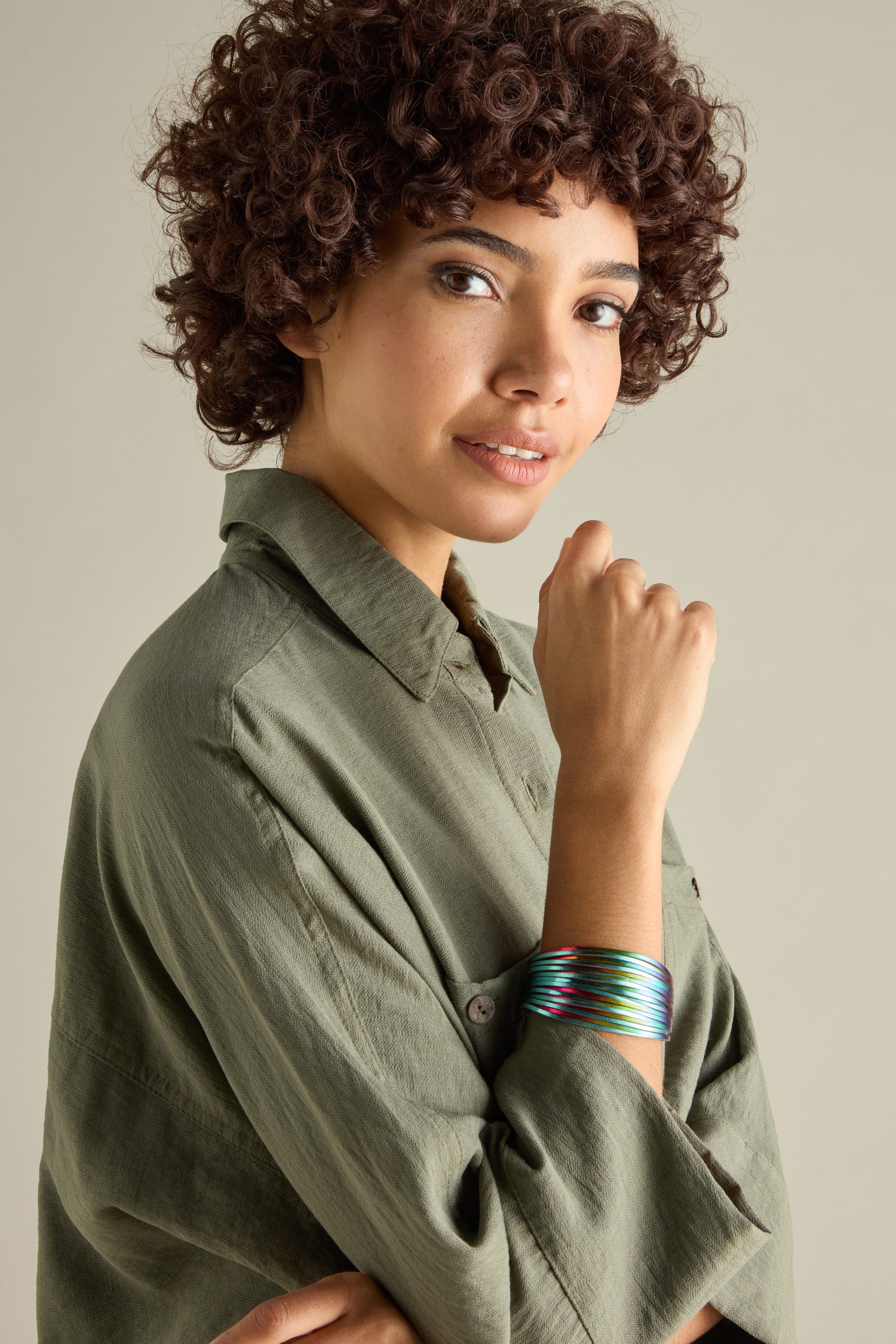 A person with curly hair smiles slightly, wearing a green shirt and colorful Woven Metallic Bracelet on their wrist.