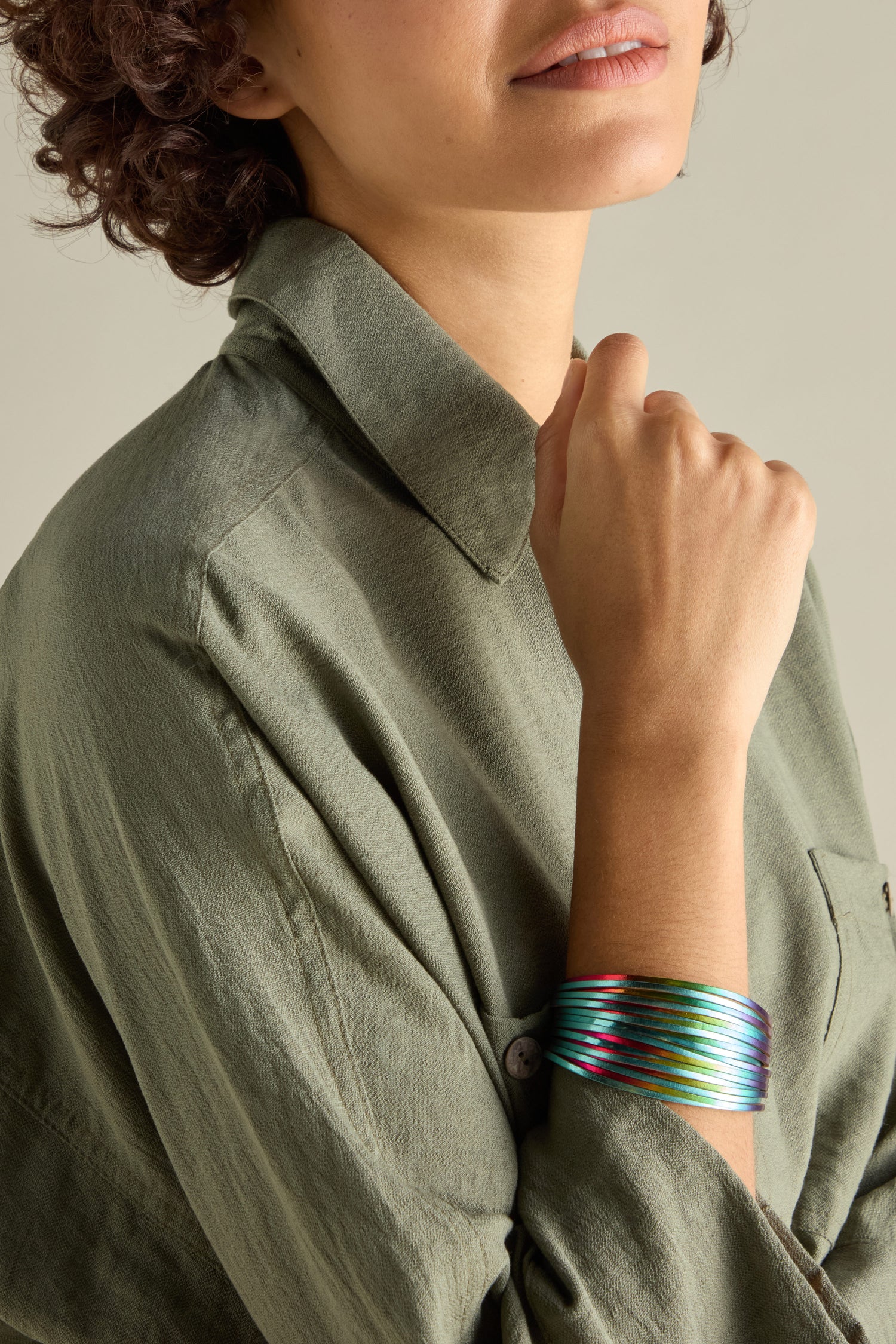 A person in a green shirt elegantly raises one hand to their face, showcasing a stack of Woven Metallic Bracelets alongside a twisted leatherette bracelet with a magnetic clasp.