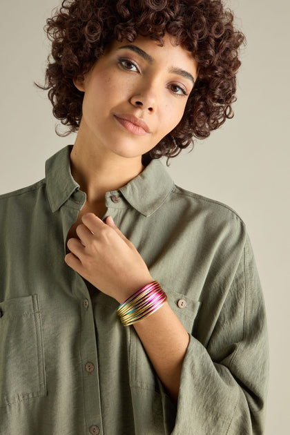 A person with curly hair in a green shirt gazes at the camera neutrally, colorful bangles and a woven metallic bracelet with a magnetic clasp adorning their wrist.