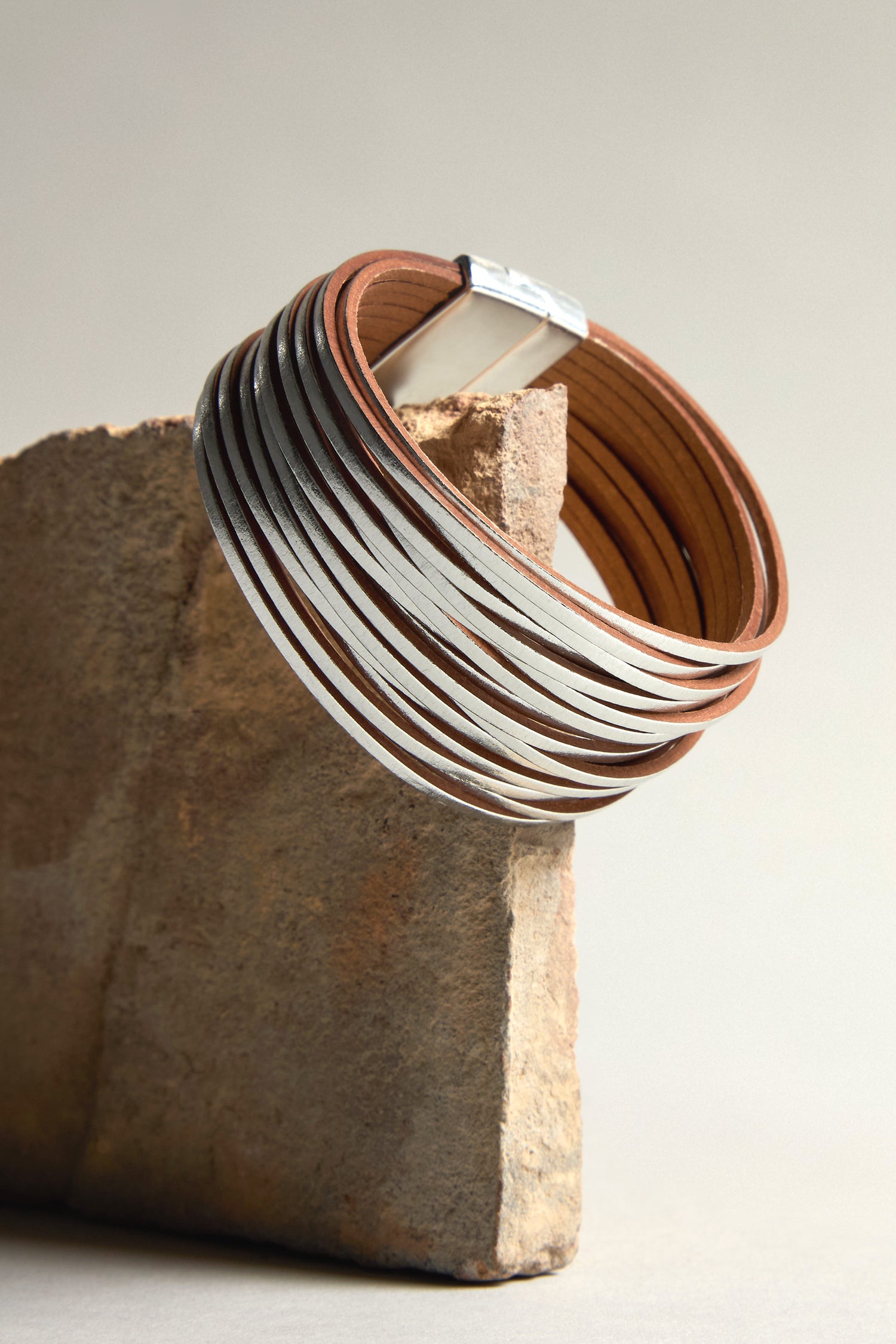 The Woven Metallic Bracelet, crafted with silver and brown leather and featuring a subtle metallic finish, is elegantly showcased on a rectangular stone slab.