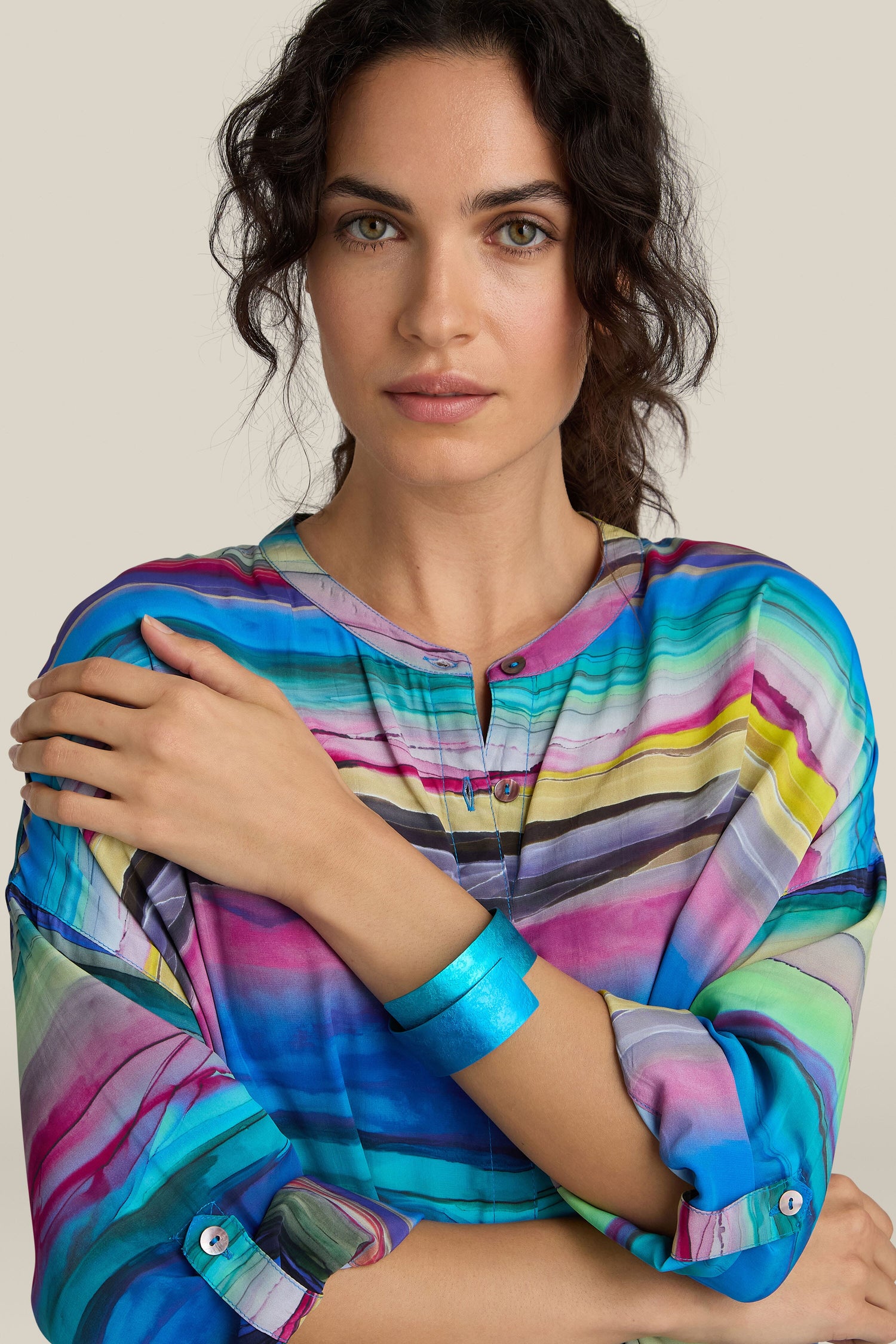 A person with curly hair wearing a colorful striped top and a handcrafted Curved Bracelet made of aluminum is standing with their arms crossed, looking directly at the camera.