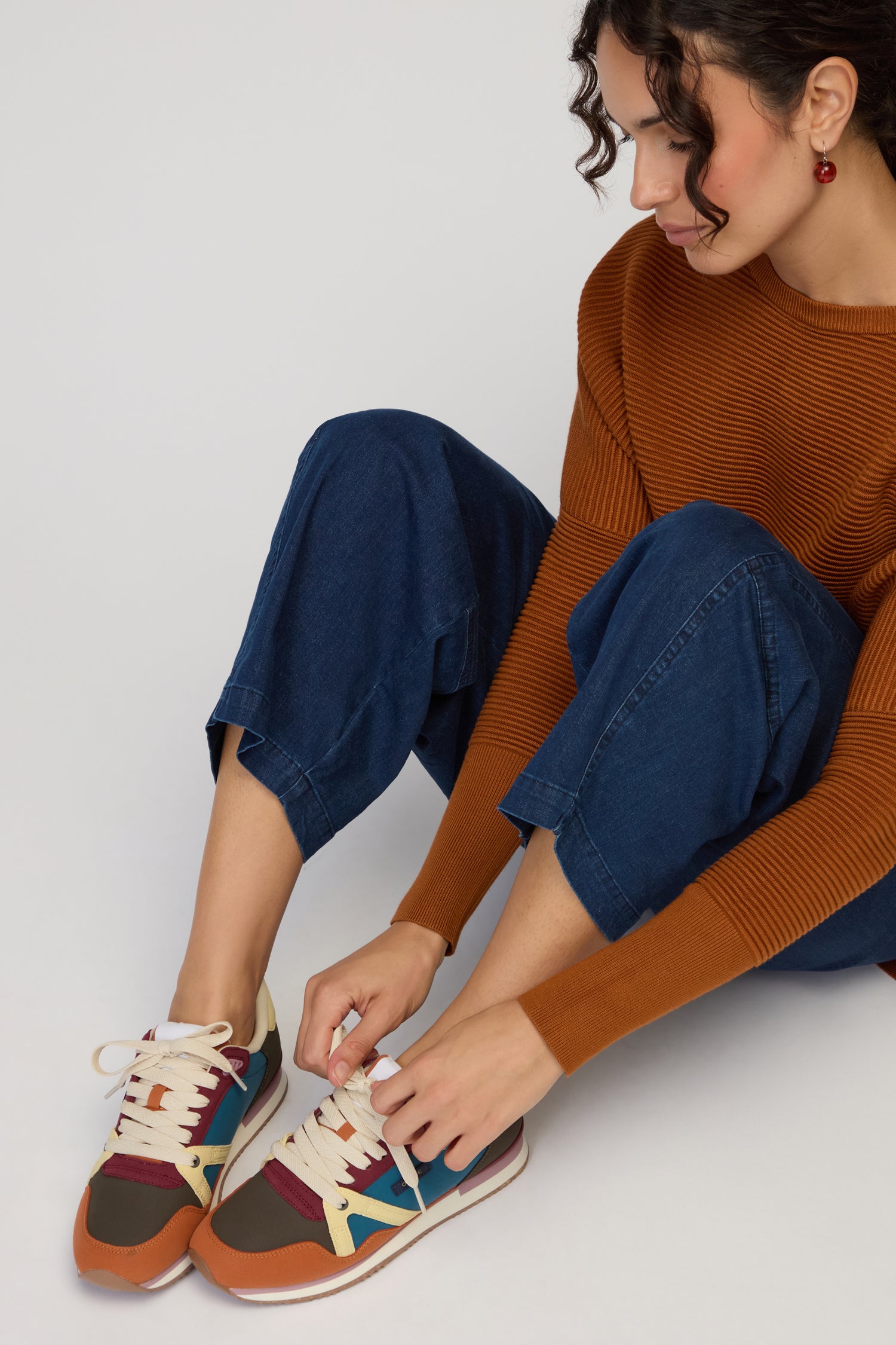 A person in a rust-colored sweater and blue jeans sits tying the laces of colourful, sustainable Colour Pop Andree Trainers, highlighting the stylish colour blocking of their sneakers.