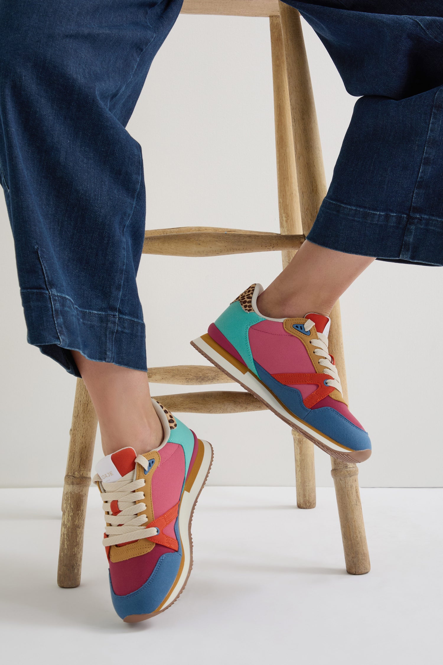 A person wearing Andree Trainers, crafted from colorful recycled mesh, and denim pants is sitting on a wooden chair.