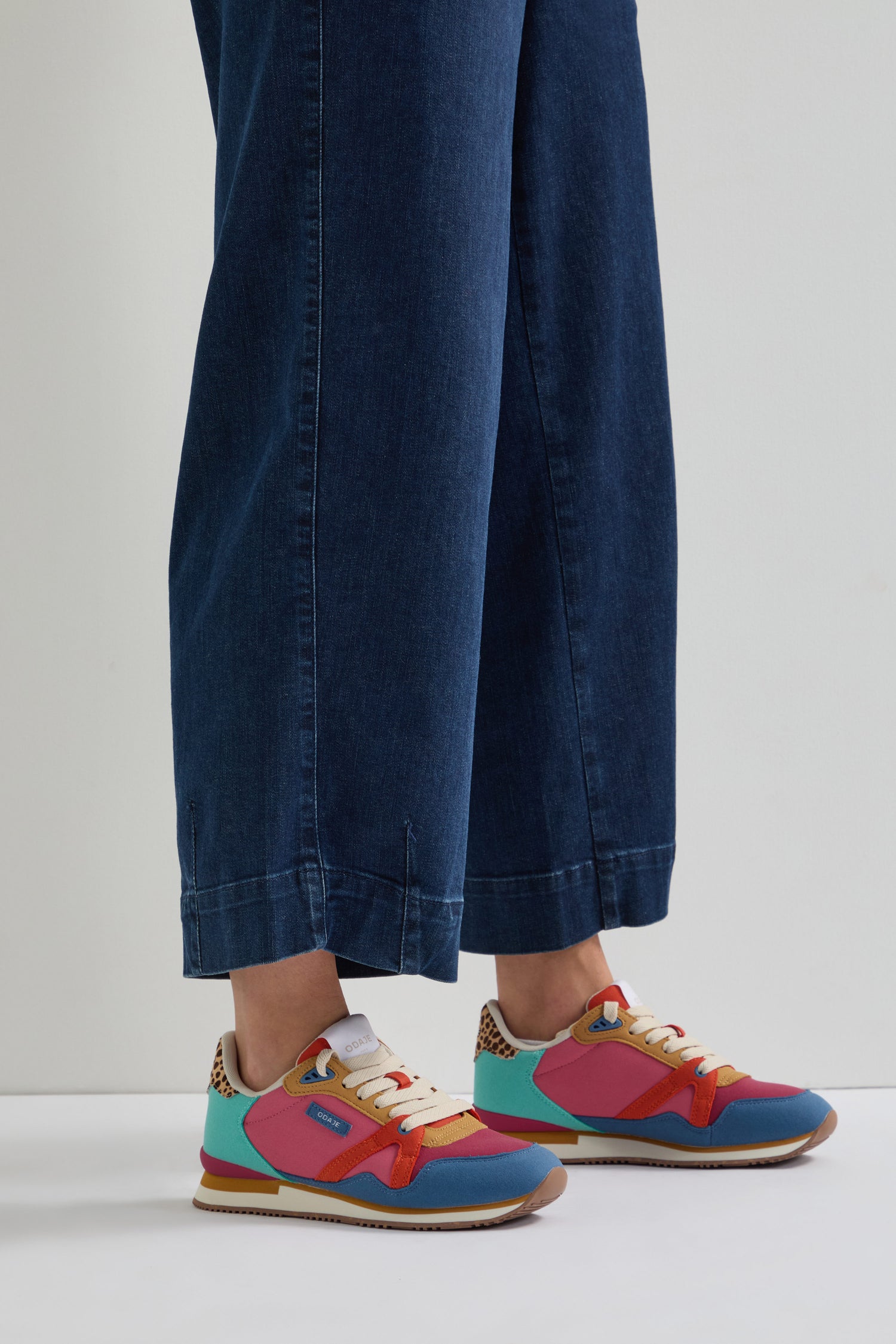 A person in blue wide-legged jeans pairs them with Andree Trainers, which feature orange, pink, and blue details and are made with recycled mesh for an eco-friendly touch.