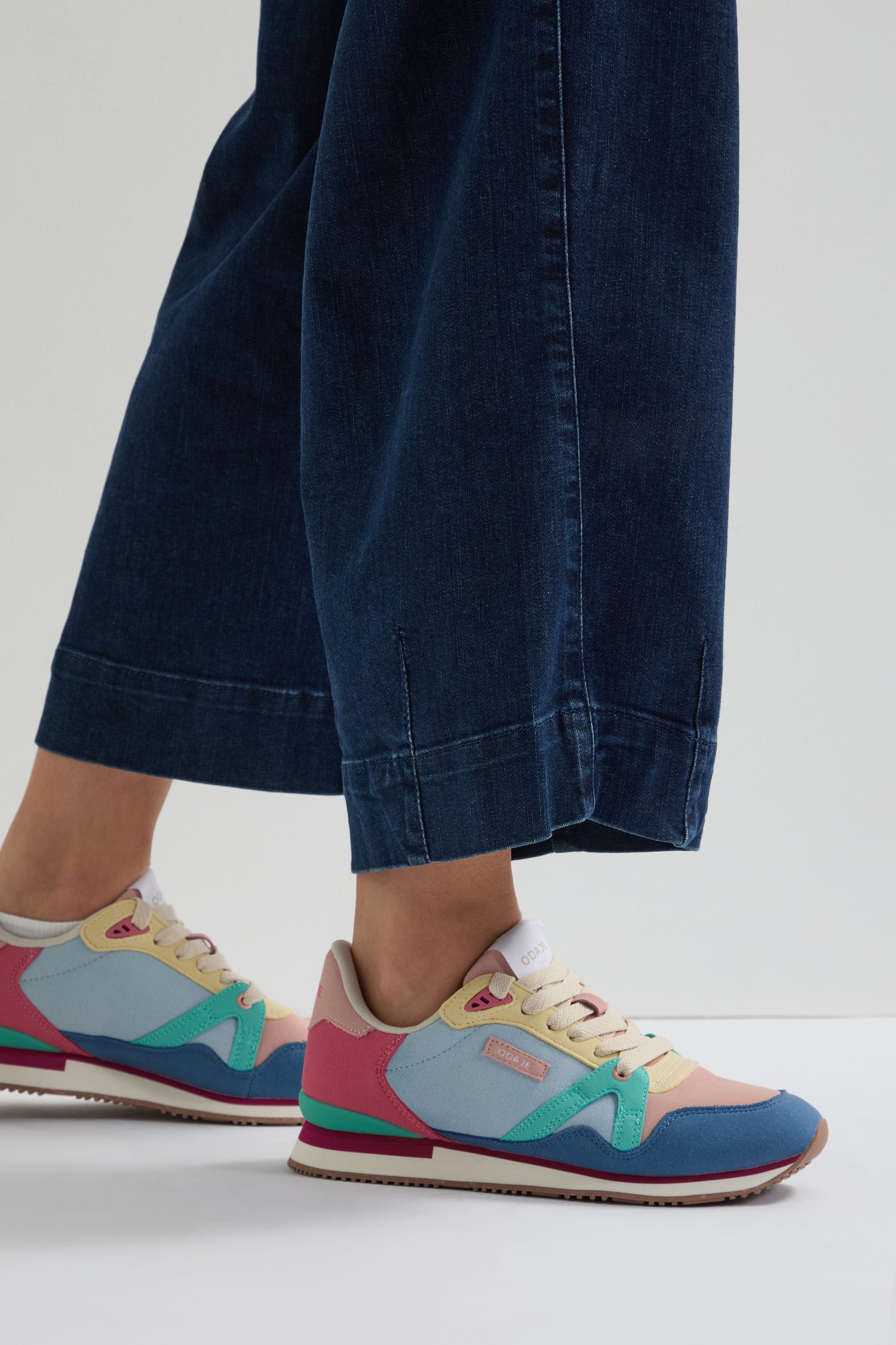 Someone wearing wide-leg blue jeans and colorful Andree Trainers, crafted with recycled mesh and vegan suede. The sneakers feature pink, blue, green, and beige accents on a gray background.