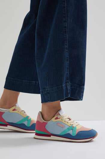 Someone wearing wide-leg blue jeans and colorful Andree Trainers, crafted with recycled mesh and vegan suede. The sneakers feature pink, blue, green, and beige accents on a gray background.