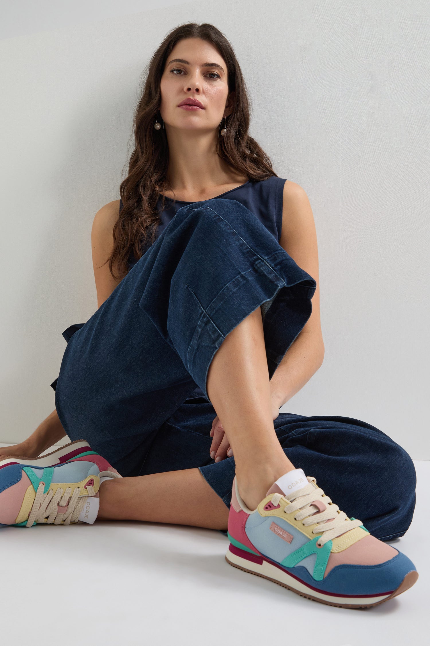 A woman in a navy jumpsuit sits on the floor with crossed legs, wearing vibrant Andree Trainers made from recycled mesh, looking towards the camera.