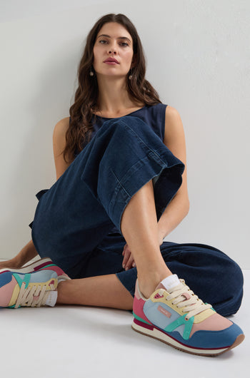 A woman in a navy jumpsuit sits on the floor with crossed legs, wearing vibrant Andree Trainers made from recycled mesh, looking towards the camera.