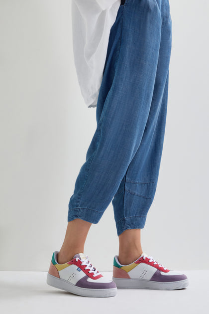 A person in blue pants and Maxence Trainers, crafted from vegan suede, stands on a white surface.