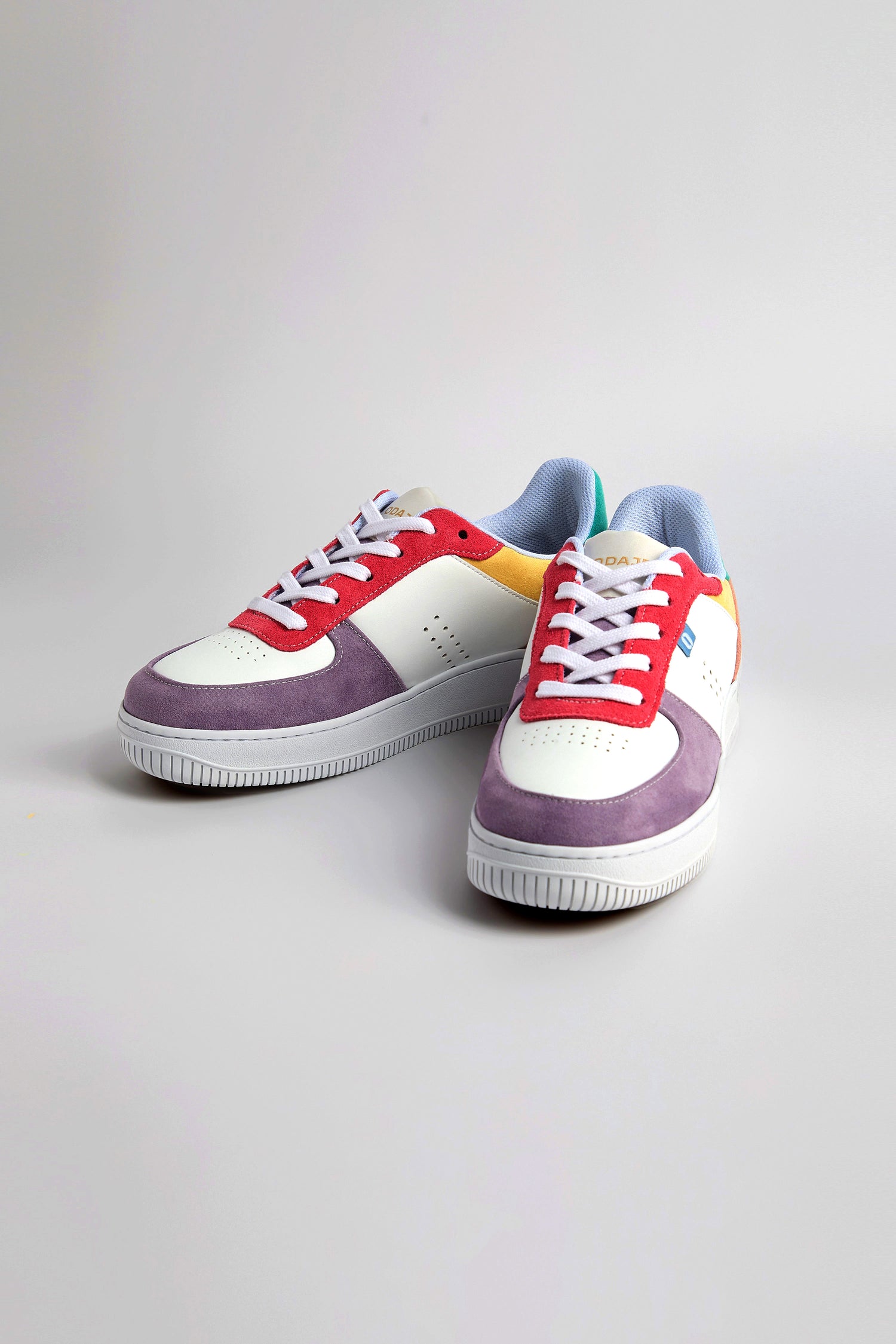 The Maxence Trainers sport colorful sneakers with red laces, purple and white panels, and blue accents, crafted from vegan suede on a light gray surface.