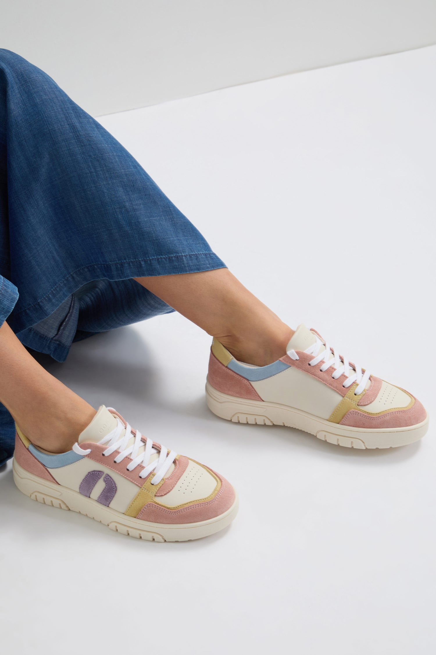 A person sits with crossed legs, displaying pastel-colored Nora Trainers and blue jeans. These sneakers highlight sustainable craftsmanship, reflecting a commitment to eco-friendly fashion.