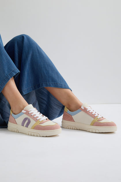 A person stands against a plain background, wearing wide-leg blue jeans and Nora Trainers made from vegan suede, featuring pastel colors and white laces.