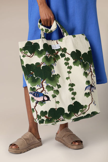 A person in a blue dress holding an Acer Canvas Bag with a leaf and bird pattern, standing in brown sandals.