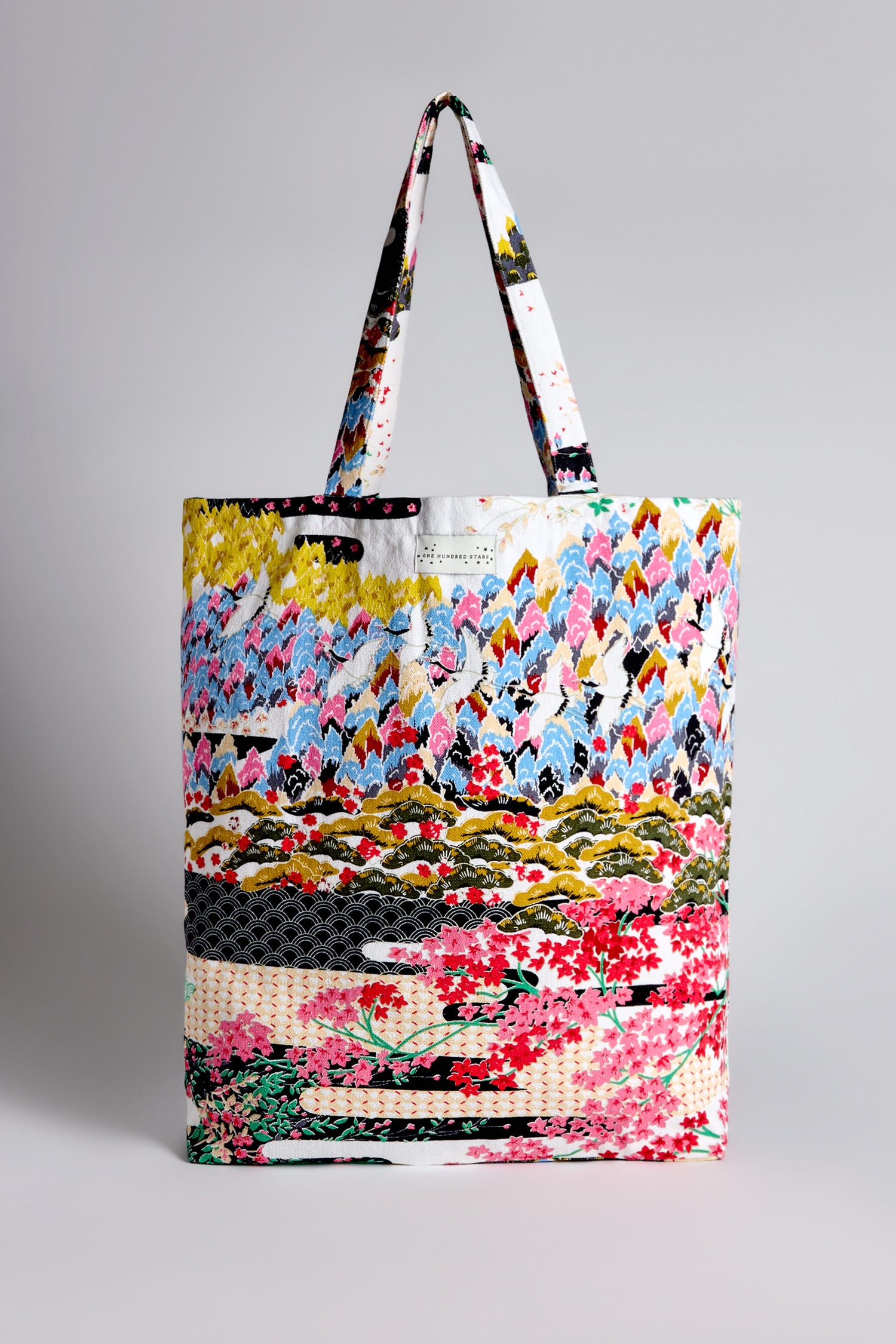 The Spring Print Canvas Bag is a vibrant patchwork tote made from sustainable cotton, showcasing floral patterns inspired by Japanese landscapes, and equipped with two handles against a simple background.