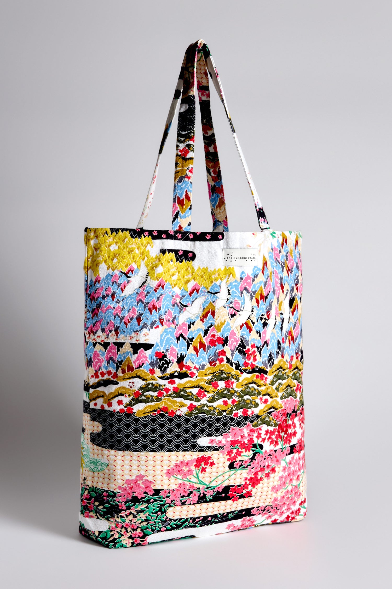 The Spring Print Canvas Bag is a colorful tote crafted from sustainable cotton, showcasing abstract floral designs inspired by Japanese landscapes, with two handles on a plain background.