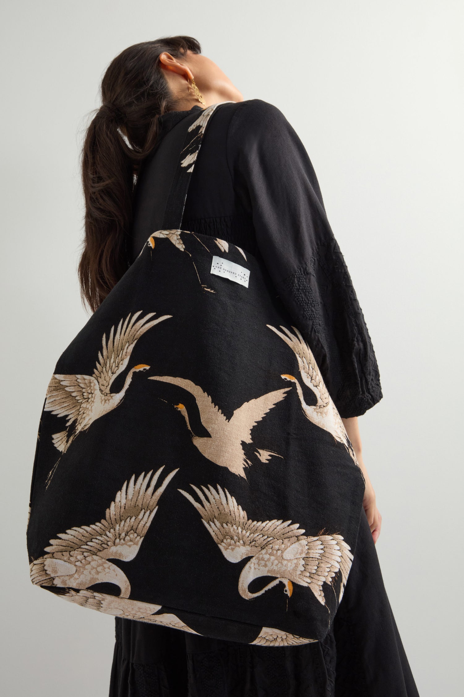 A dark-haired woman in a black dress holds the eco-friendly Stork Canvas Bag, made from sustainable cotton and featuring a striking crane design.