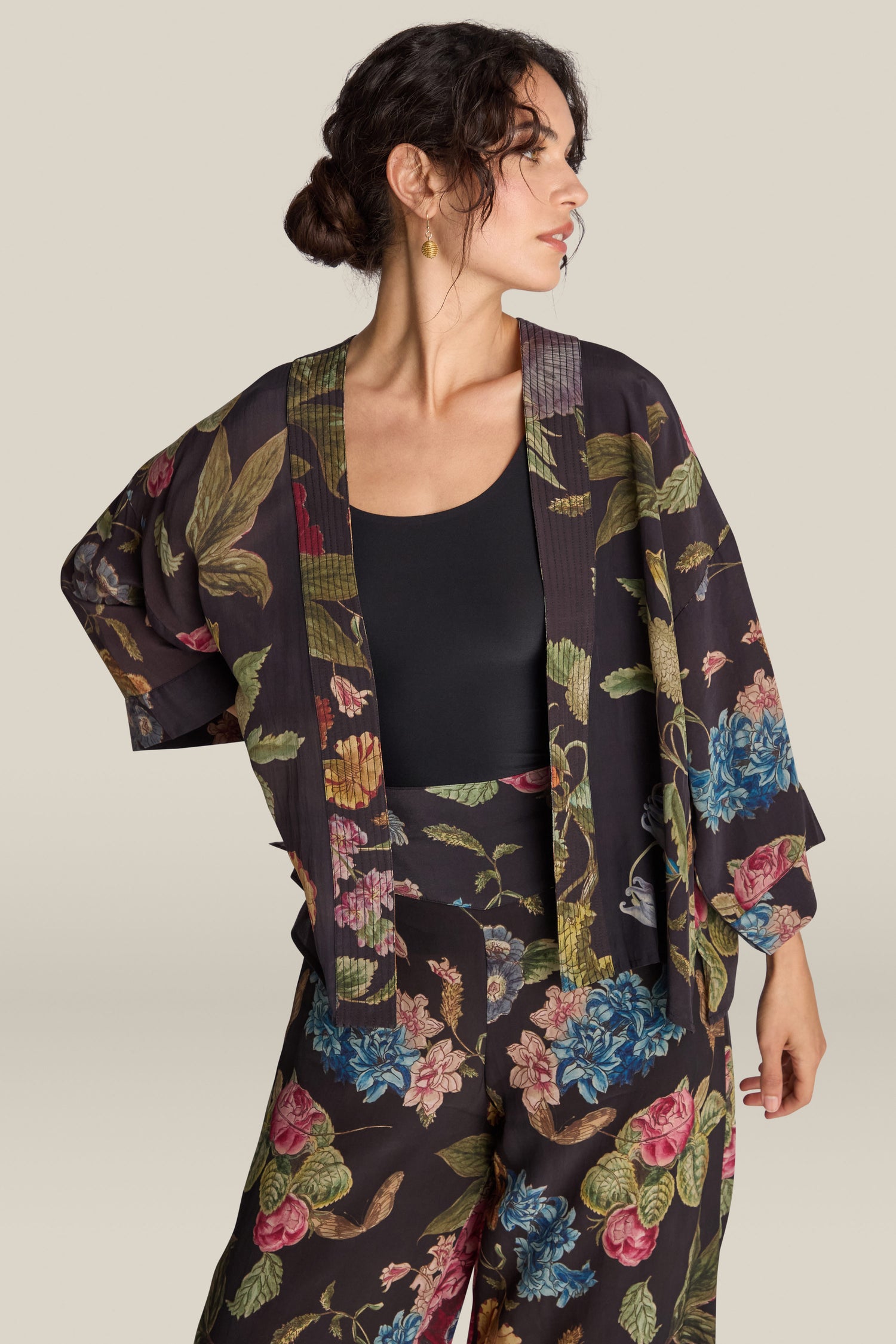 A woman with dark hair styled in a bun is wearing a black top paired with matching floral print kimono jacket and pants. She exudes elegance, accessorized with the luxurious Primula Crepe Kimono, as she looks to the side, one hand confidently placed on her hip.