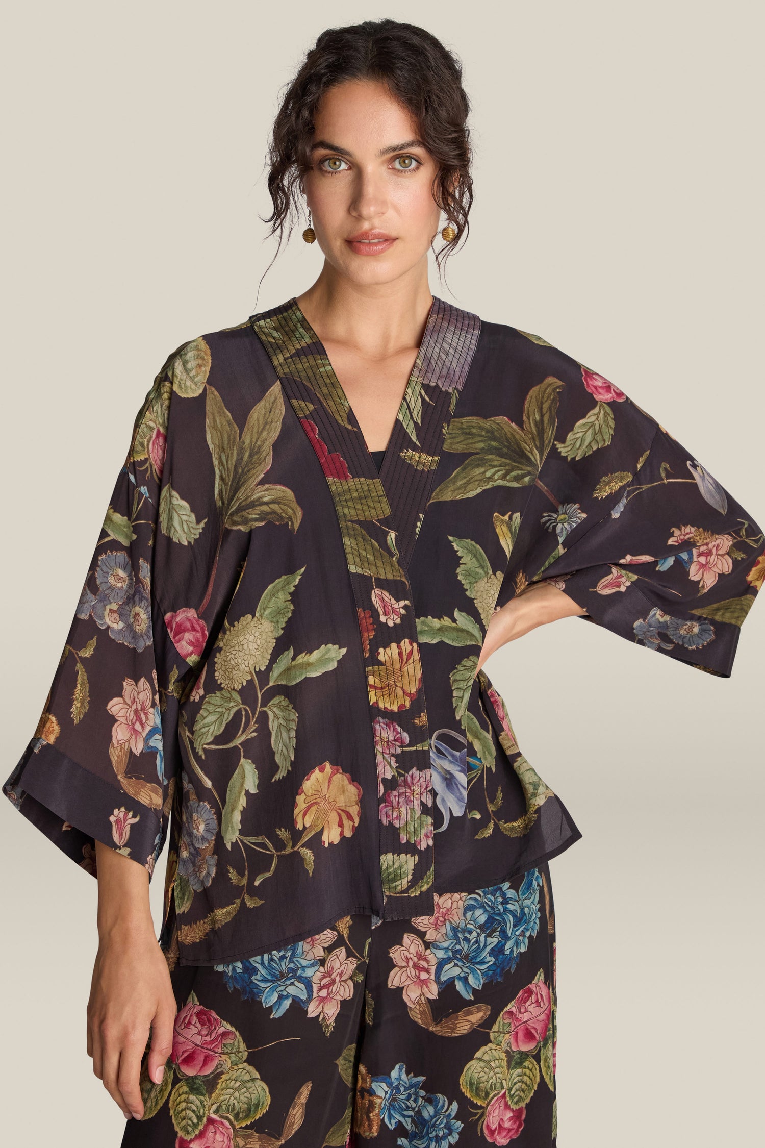 A woman wearing the Primula Crepe Kimono, a dark floral print top with matching pants, stands with one hand on her hip, looking directly at the camera.