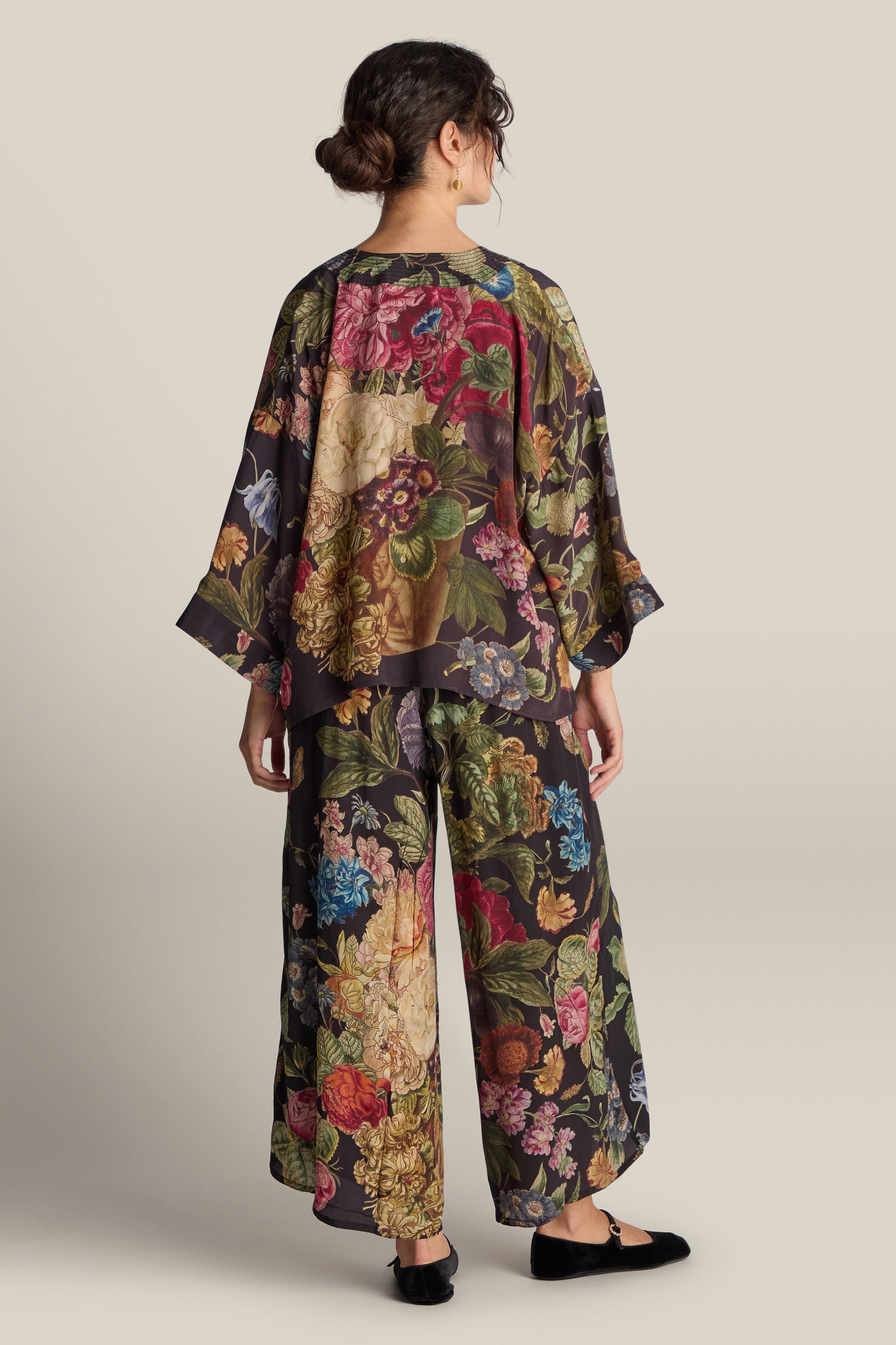 A person stands with their back to the camera, dressed in the Primula Crepe Kimono, which features a loose top and wide-legged pants, complemented by black shoes. Their hair is tied back against a plain beige background.