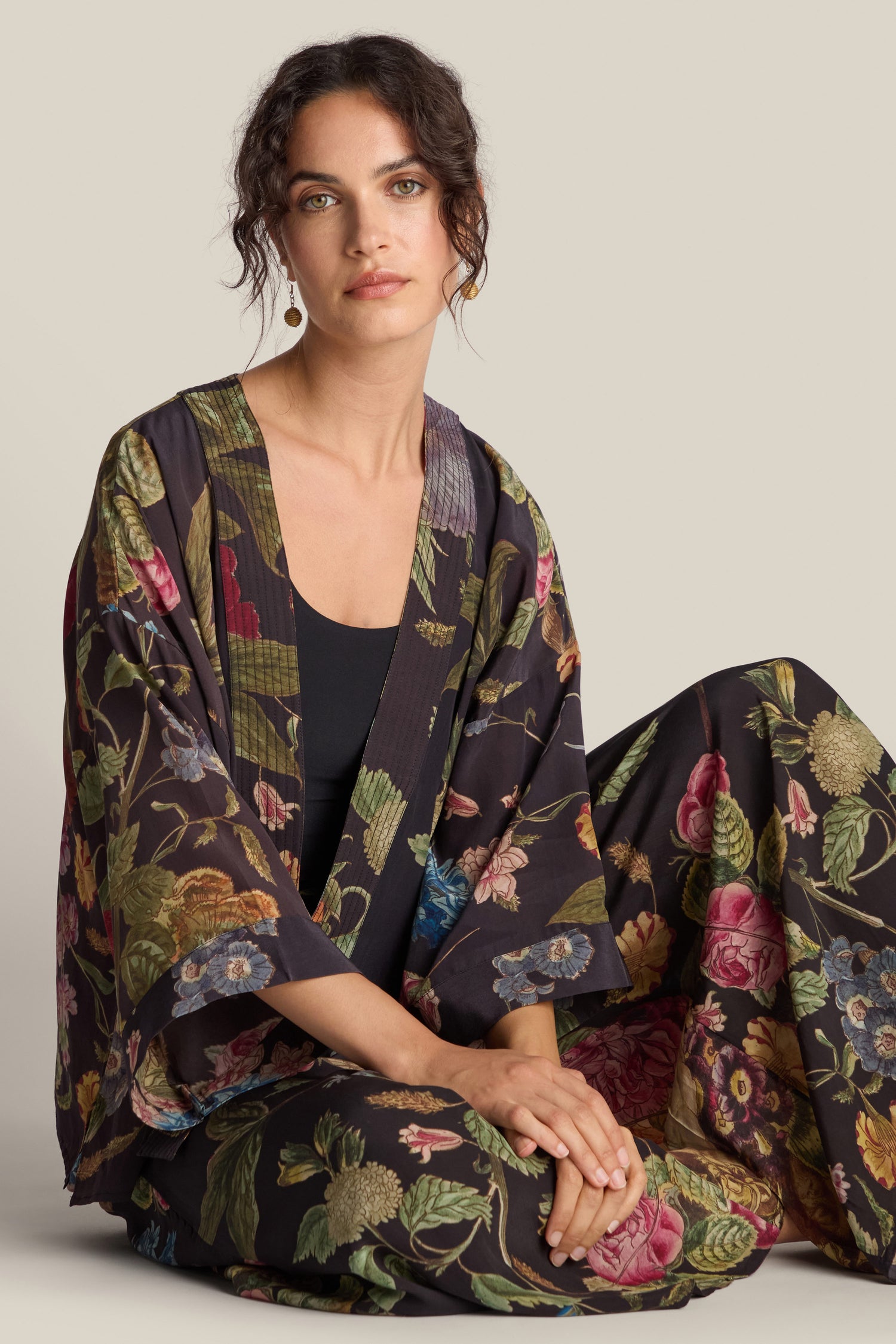 A person sits cross-legged, draped in the Primula Crepe Kimono from the One Hundred Stars collection, with a neutral background.