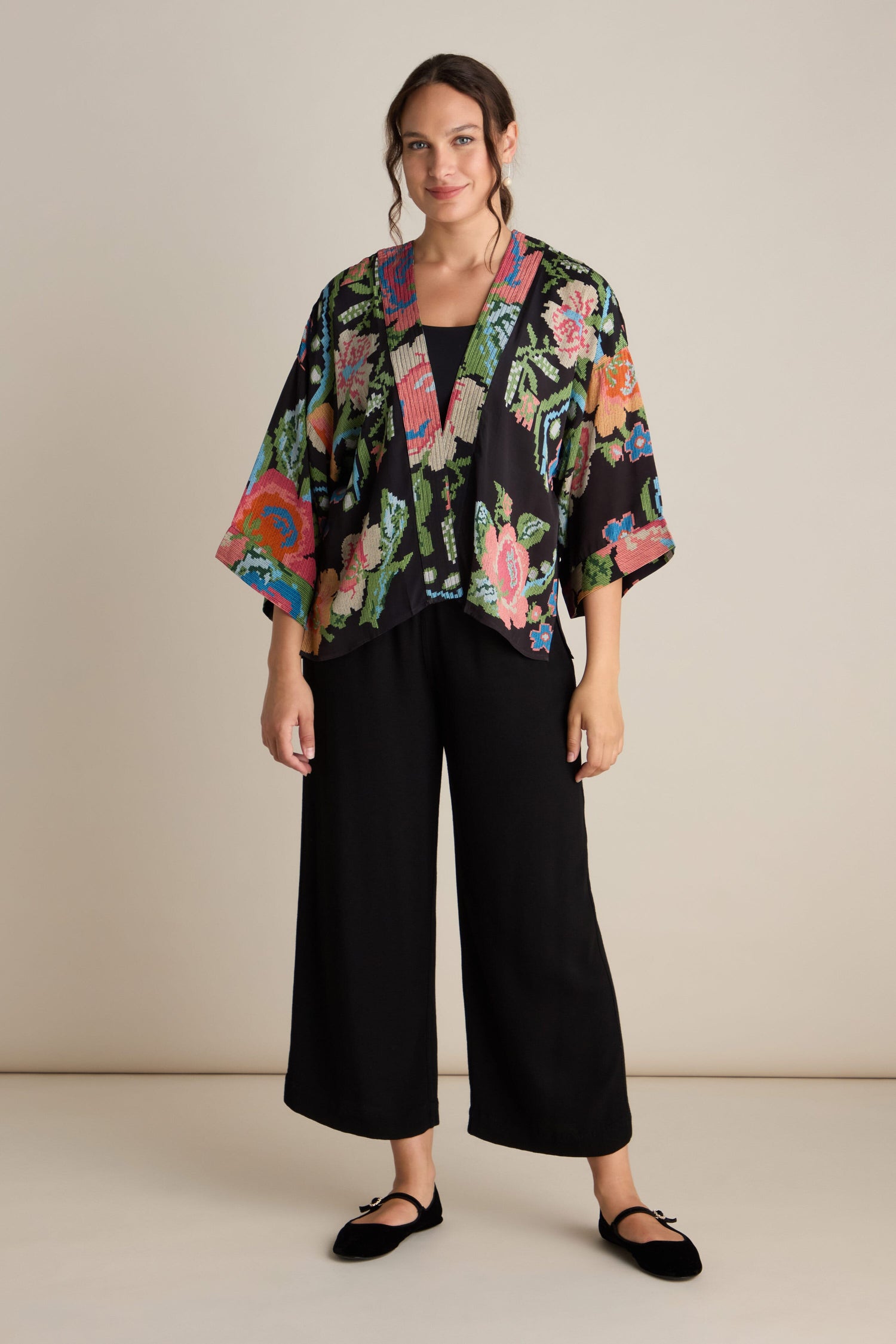 A person stands against a plain backdrop, wearing black wide-leg pants and the Woven Flower Crepe Kimono, which features a vibrant floral motif, along with black flats.