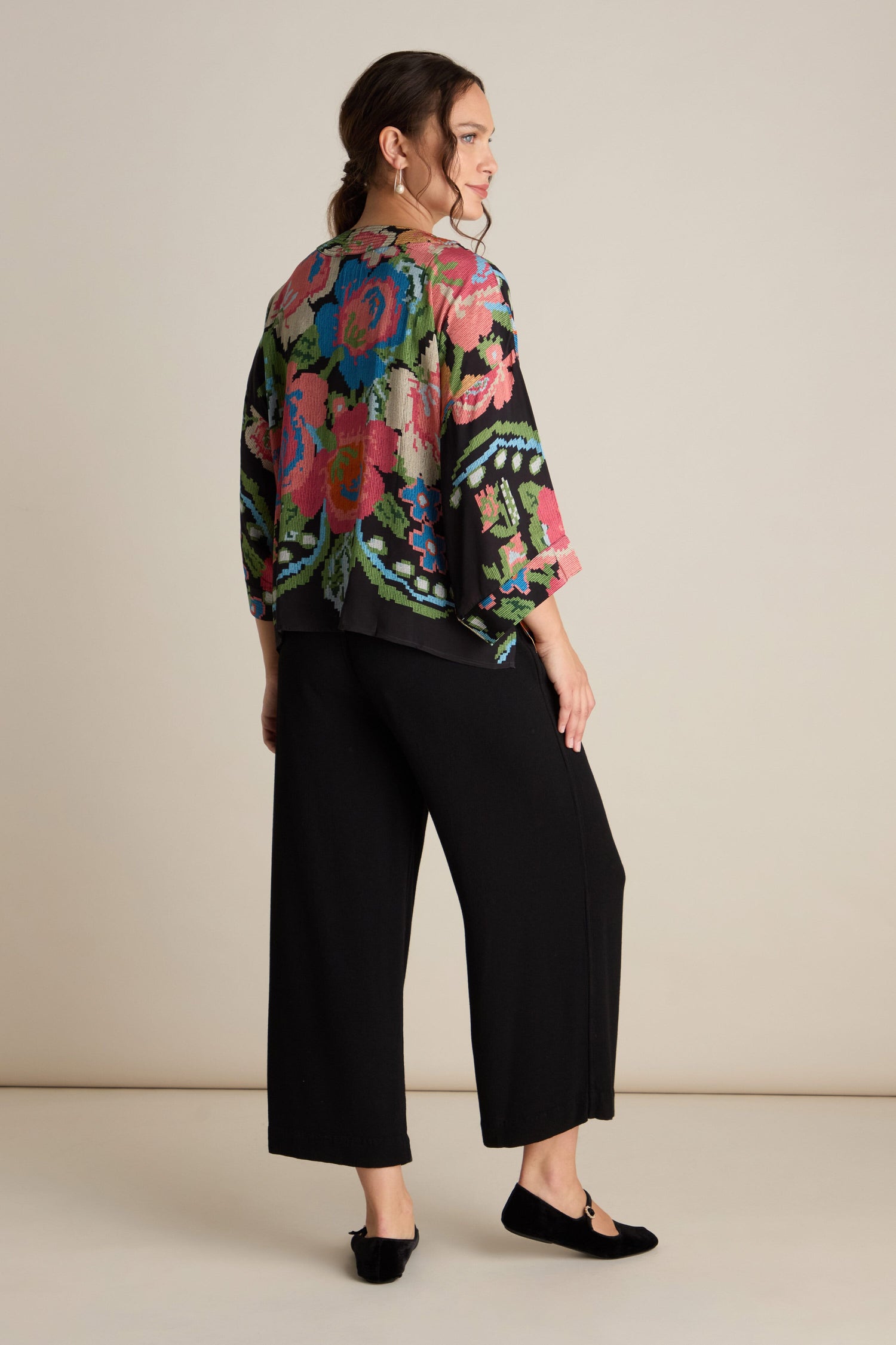 A person stands facing away in a Woven Flower Crepe Kimono, featuring a vibrant floral motif, complemented by black wide-leg pants and matching shoes, set against a plain background.