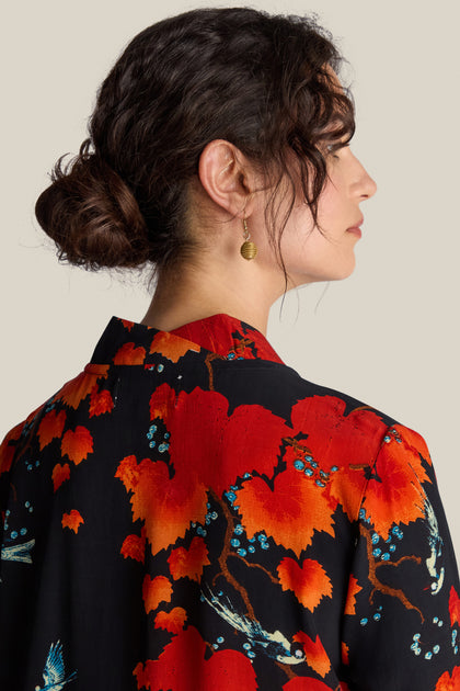 A woman with her hair in a bun is shown from the side. She is wearing the Acer Print Asymmetric Dress, which features a black garment with a red floral pattern and gold dangling earrings, reminiscent of Chinese art.