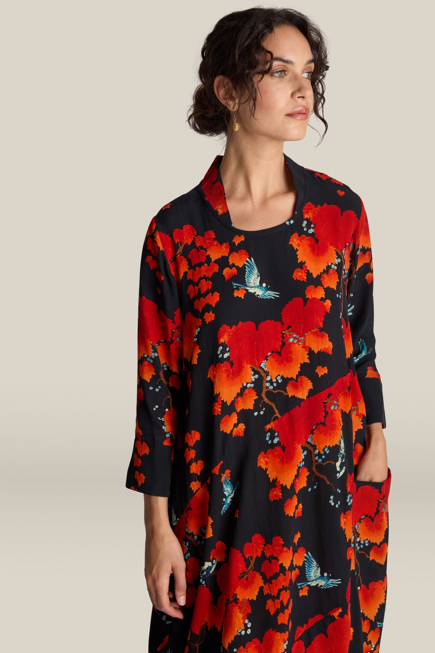 A woman with short curly hair wears an Acer Print Asymmetric Dress, showcasing a vibrant red and blue nature-themed pattern adorned with birds and trees. Inspired by Chinese art, she stands with her hand in her pocket, gazing to the side.