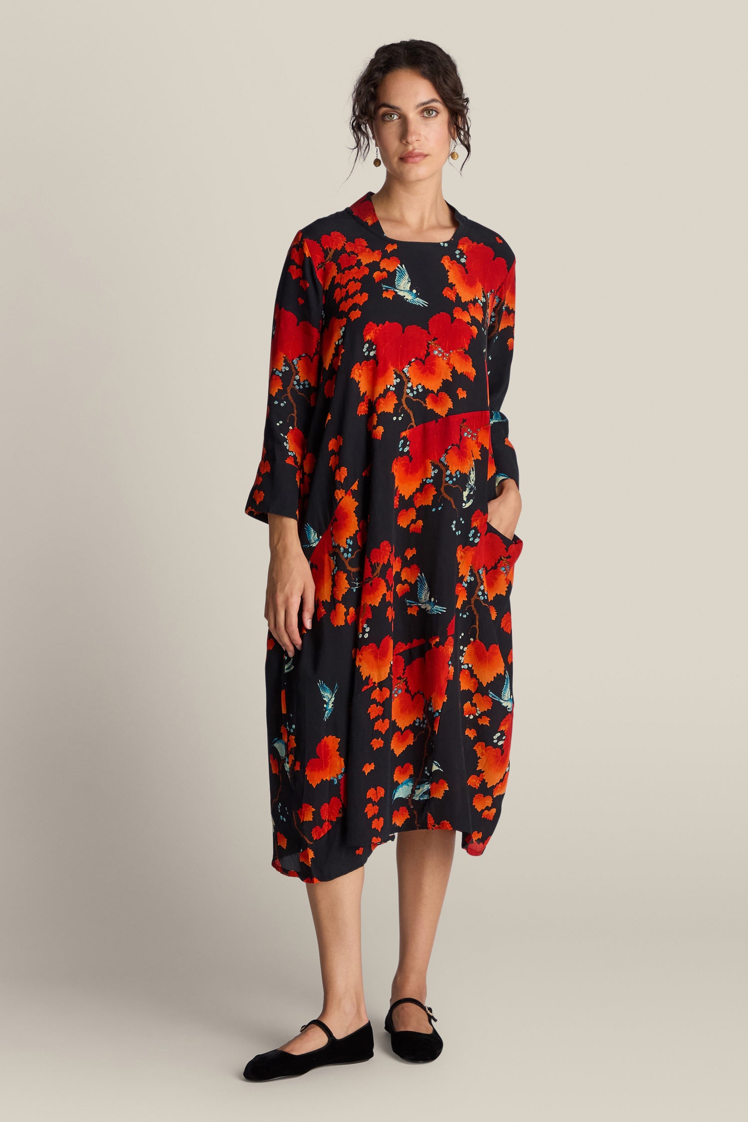 A woman wearing the Acer Print Asymmetric Dress, inspired by Chinese art, stands against a plain background. The black dress features a striking red floral pattern. With one hand in her pocket, she gazes at the camera.