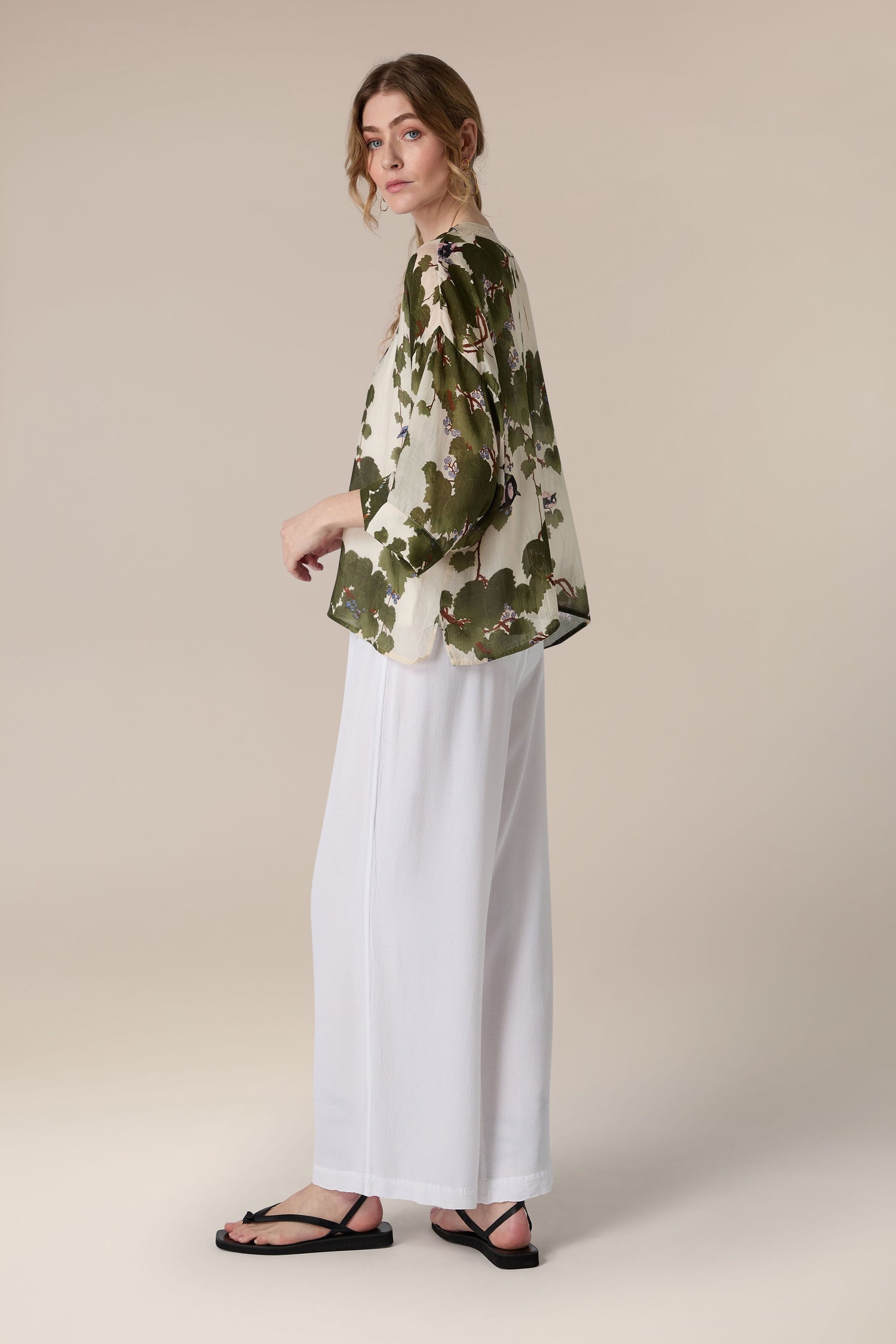 A woman posing in profile wearing a floral blouse and white trousers against a backdrop featuring an Acer Kimono.