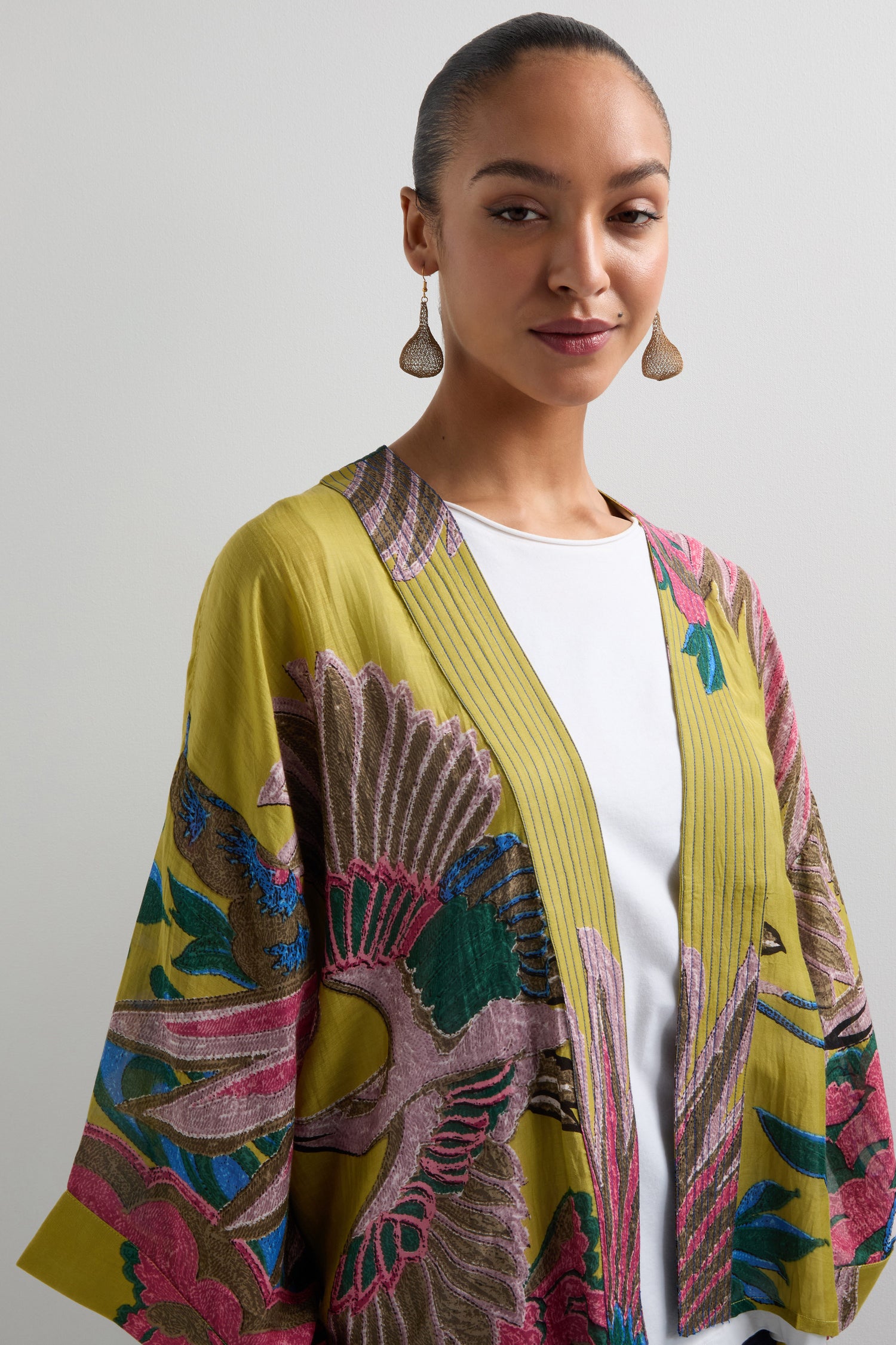 Against a plain background, an individual with tied-back hair and drop earrings wears a vibrant Crane Print Kimono by One Hundred Stars over a white top.