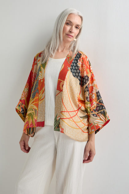 A woman with long gray hair wears the Fans Print Kimono over a white textured outfit, standing against a plain white wall.