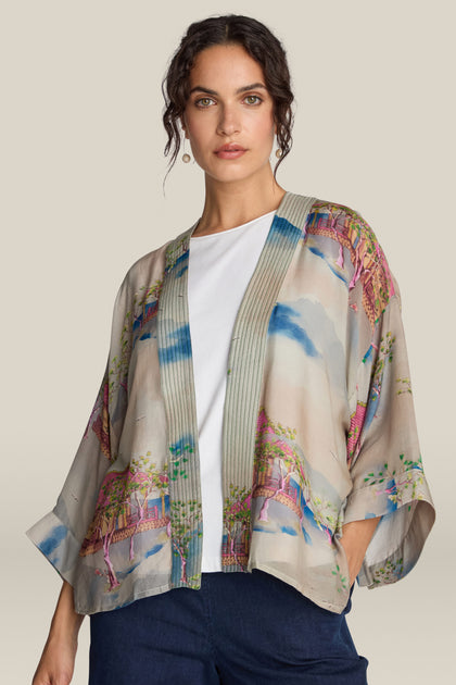 A woman wears the "Landscape Kimono," a short jacket patterned like Japanese landscape watercolors, over a white shirt and dark blue jeans, standing against a plain background.