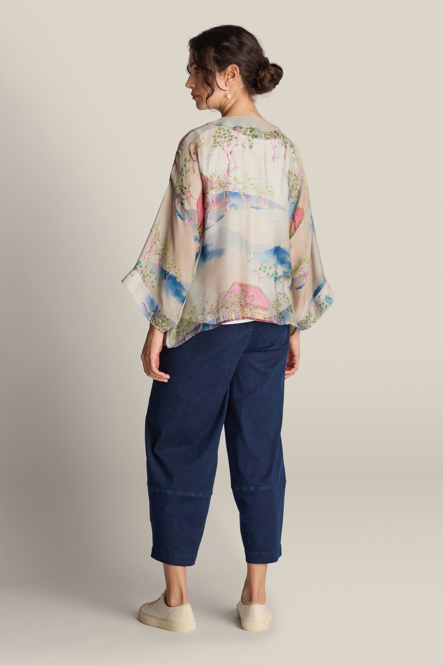 A person with hair tied back stands facing away, wearing the Landscape Kimono blouse, which features colorful patterns reminiscent of Japanese landscape watercolours. They pair it with blue cropped pants and beige shoes against a plain background.