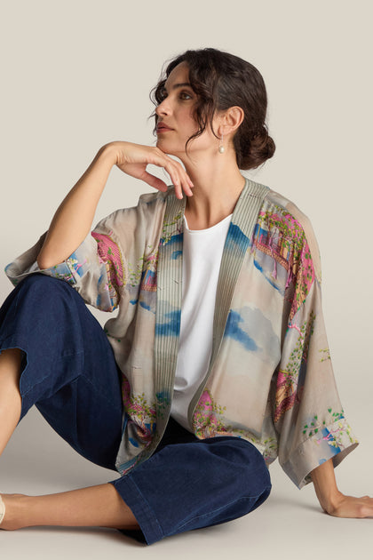 A woman with dark hair sits on the floor, leaning her head on her hand. She is wearing a short Landscape Kimono adorned with Japanese landscape watercolors, a white top, and dark pants.