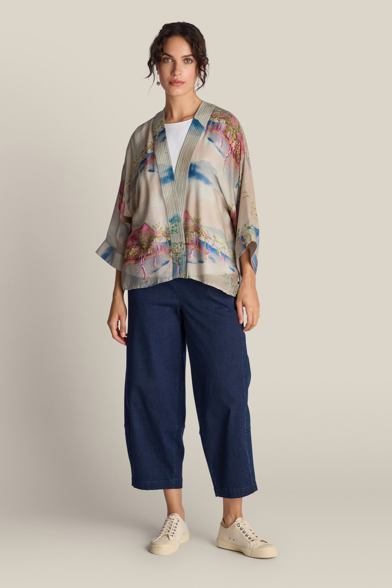 A person stands against a neutral background, wearing the Landscape Kimono adorned with Japanese landscape watercolours, paired with dark blue wide-legged pants and white sneakers.