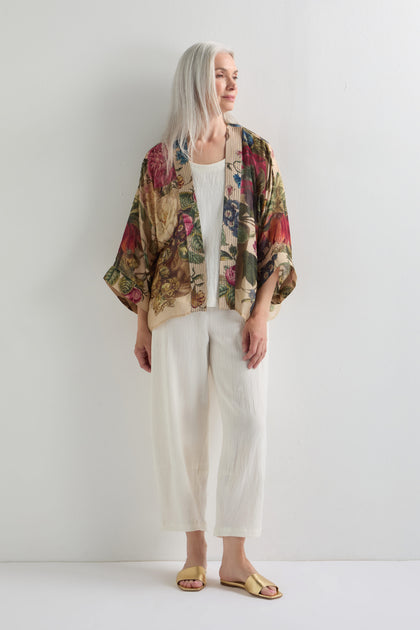 A woman with long gray hair exudes artistic flair in the bold Primula Kimono over a white outfit and gold sandals, standing against a plain white wall.