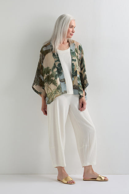 A woman with long gray hair models the Rivers and Trees Print Kimono over a cream top and pants with yellow sandals, standing against a plain white wall.