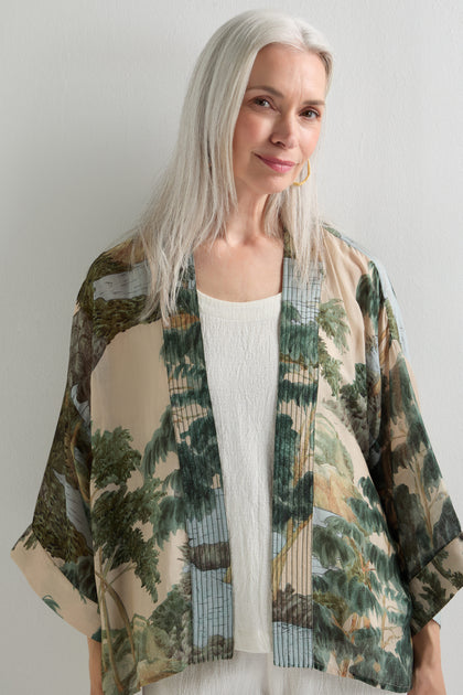 An older woman with long gray hair stands against a plain wall, wearing the Rivers and Trees Print Kimono as a summer cover-up over a white top.
