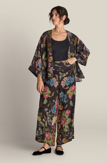 A person stands wearing a black floral kimono and matching Primula Palazzo Trouser over a black top. Their hair is styled back, and they are wearing black flat shoes. The background is plain beige.