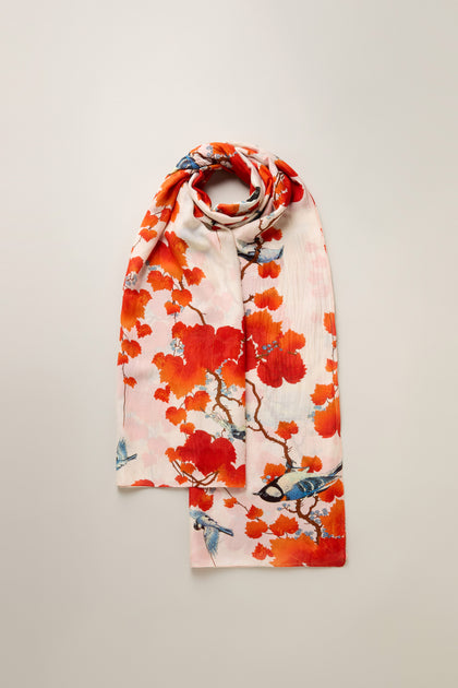 The Acer Print Scarf is a folded pink scarf adorned with vibrant orange floral and bird patterns on a light background, crafted from a silky modal-cupro blend.