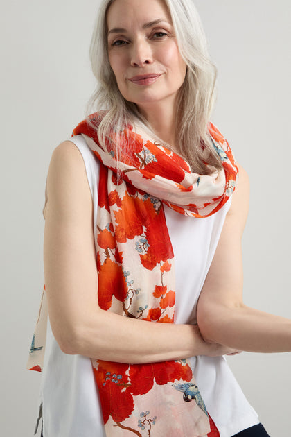 A woman with long gray hair wears a white sleeveless top, her look enhanced by the red and white Acer Print Scarf, crafted from a silky modal-cupro blend, as she stands against a plain background.