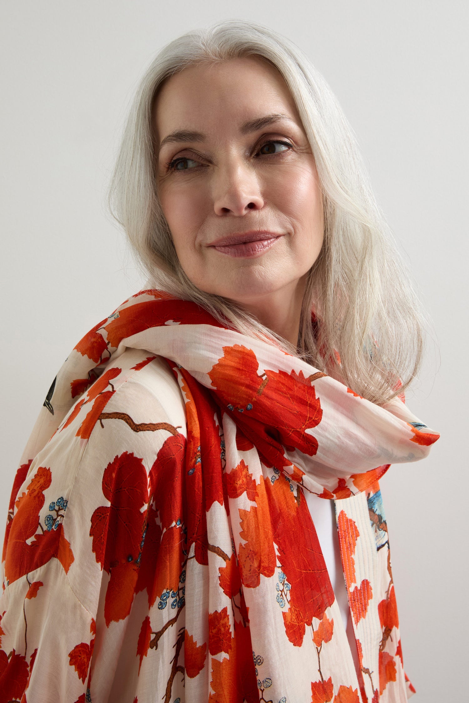 A person with long white hair wears a floral-patterned top and an Acer Print Scarf in modal-cupro, gazing sideways against a neutral backdrop.