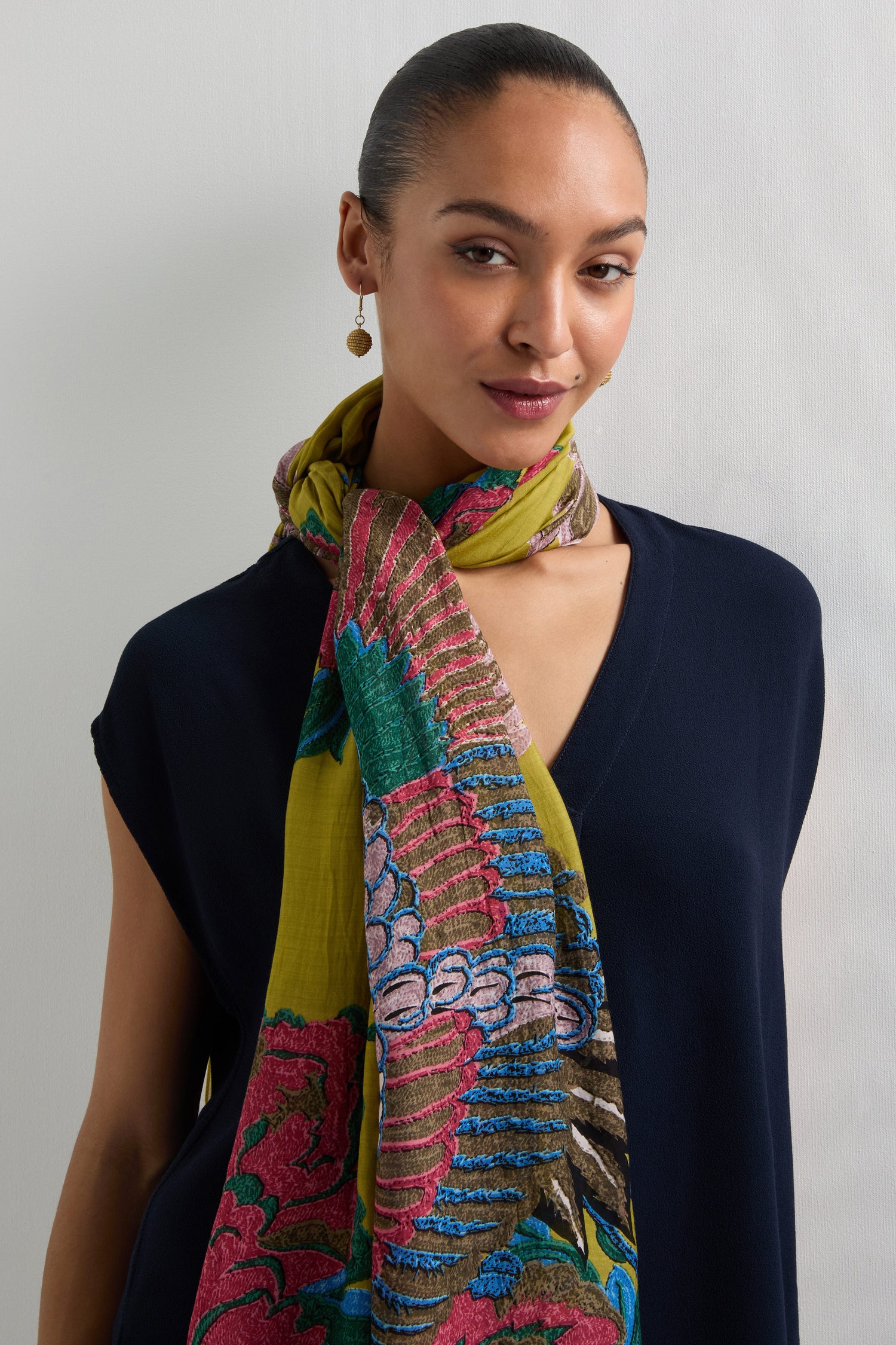 A person in a navy top stands against a light background, wearing the Crane Print Scarf adorned with colorful floral and abstract patterns. This elegant accessory enhances their outfit with vibrant flair, reminiscent of elaborate kimonos.