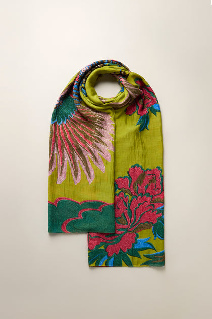 A Crane Print Scarf in green features vibrant floral patterns in pink, red, and teal, evoking intricate kimono designs, beautifully folded against a simple backdrop for contrast.