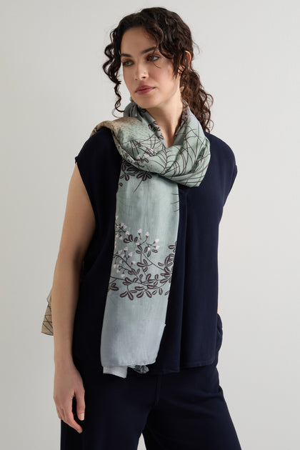 A person in a navy outfit and the Dunes Print Scarf with an intricate hand-drawn design looks sideways against a plain backdrop. The lightweight, breathable fabric of the scarf adds elegance to their ensemble.