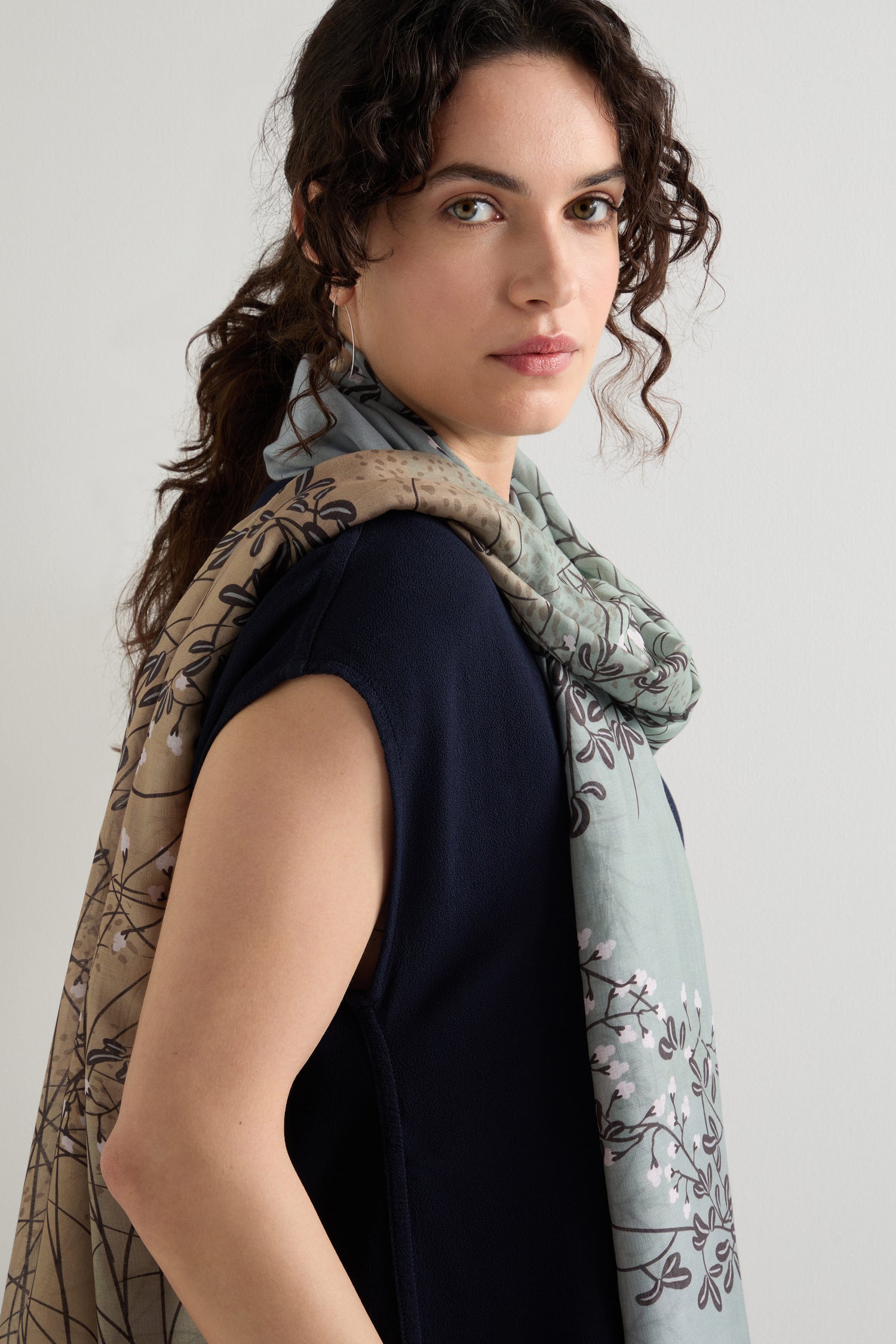 A curly-haired person in a navy sleeveless top and a lightweight Dunes Print Scarf poses against a plain backdrop, looking at the camera.